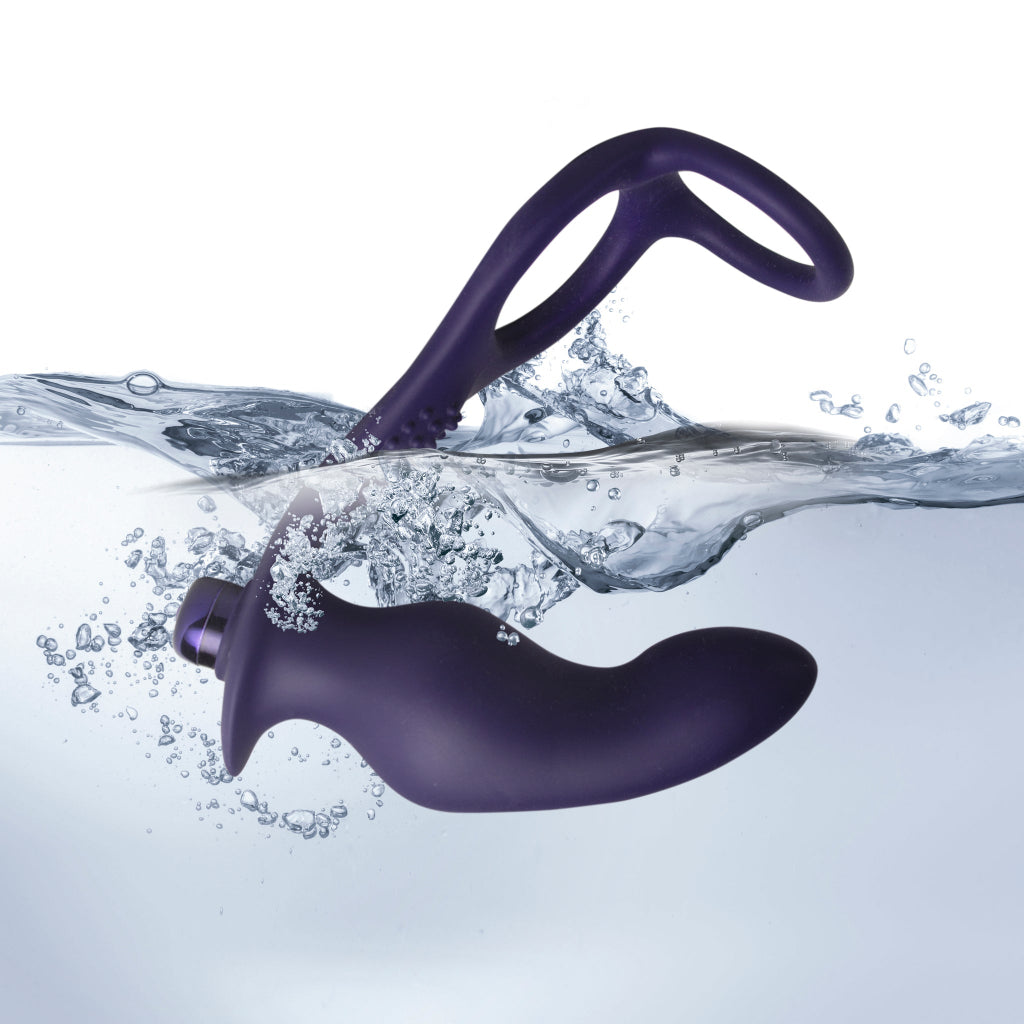 An action photograph of a dark purple silicone prostate massager partially submerged in clear, splashing water against a white background. The wavy water surface splits the device, with the smooth, curved main bulbous arm resting fully underwater amidst small air bubbles. The textured lower section and its open dual-ring loop handle extend out above the waterline, highlighting the toy's waterproof design