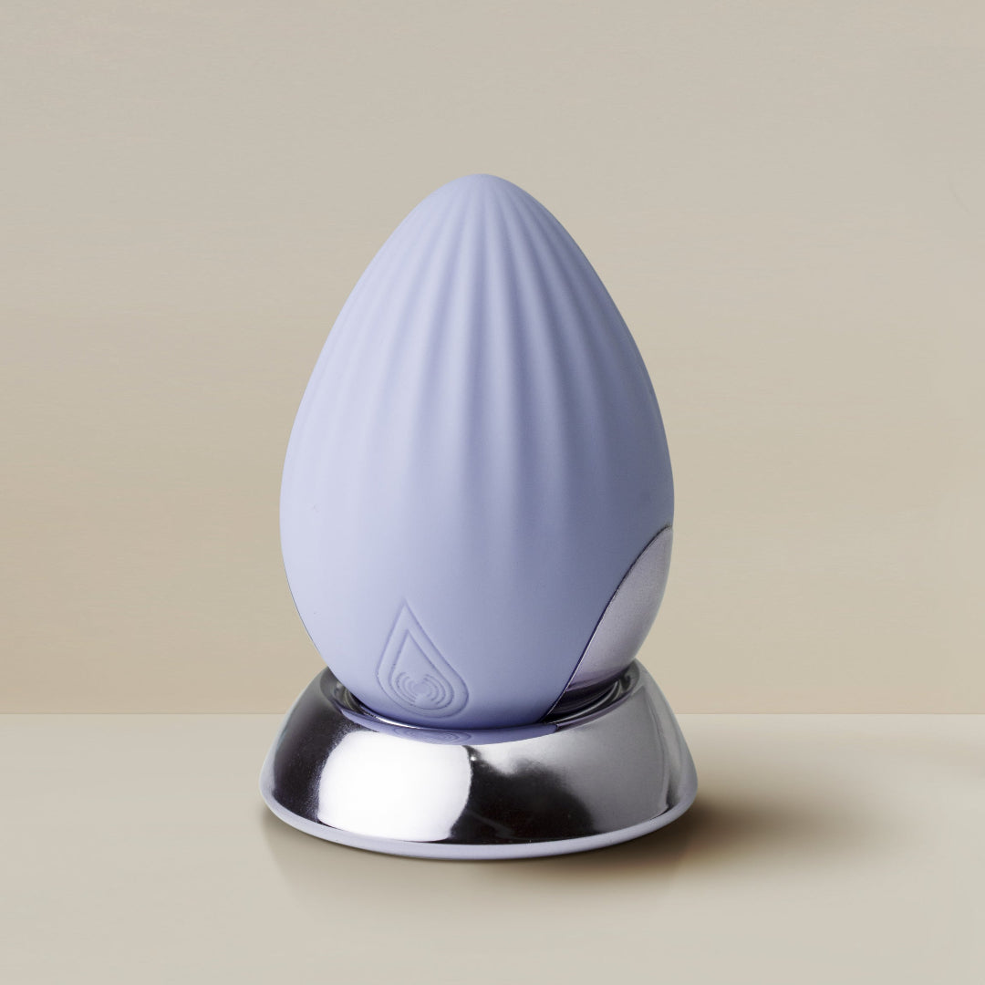 A light lavender silicone teardrop-shaped NIYA N4 egg vibrator with a ribbed texture, resting on a reflective silver metallic charging base against a warm beige background.