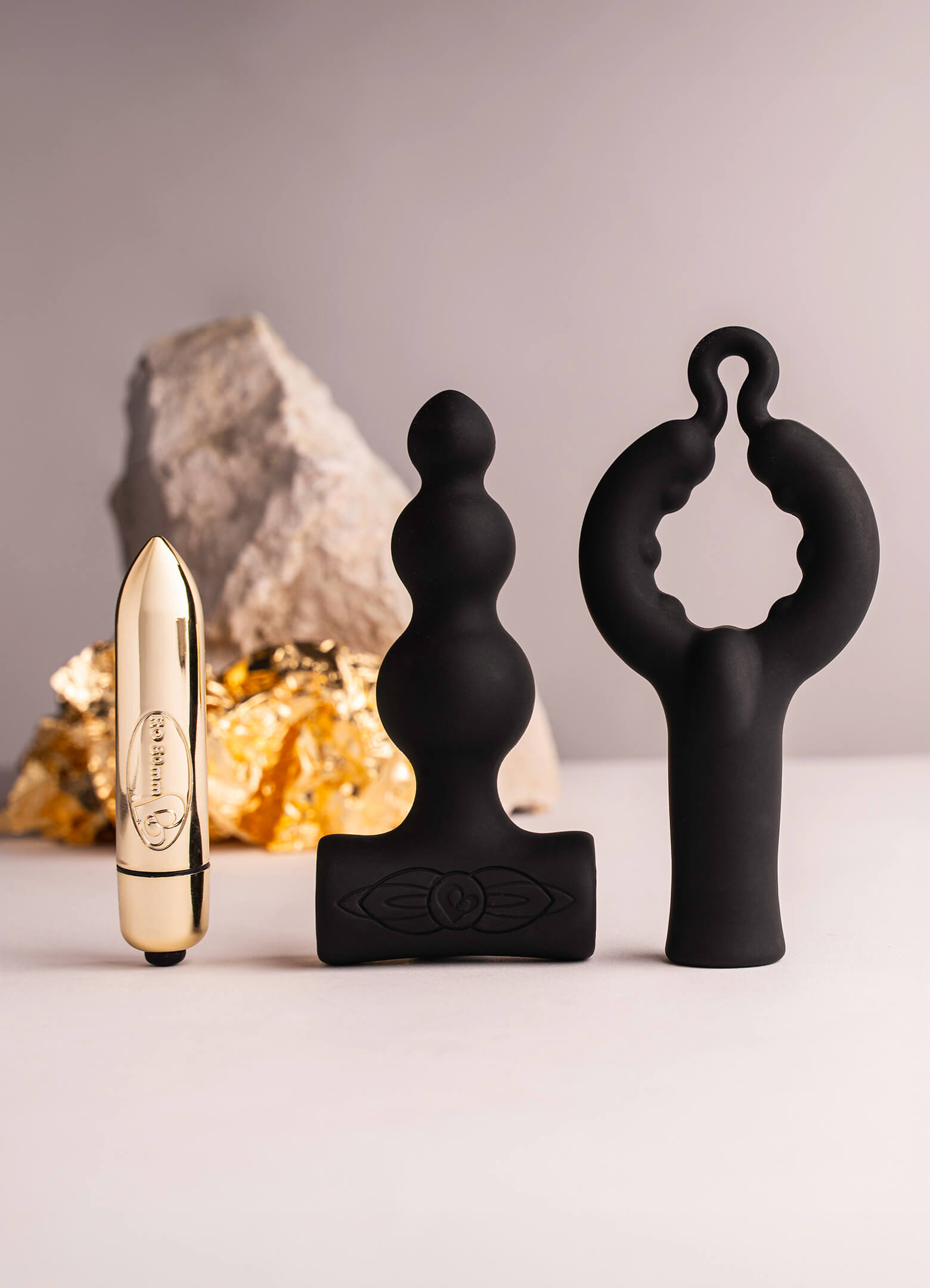 Small gold bullet vibrator next to a black bubbled silicone butt plug attachment and a black silicone cock ring attachment.