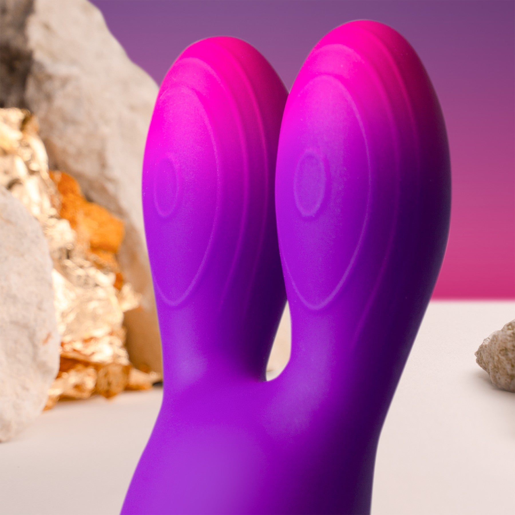 A close-up of a purple and pink silicone rabbit-style dual-stimulation Glo-Girl Bunny vibrator with a matte finish, set against a background of blurred gold and white crystals and a purple-to-pink gradient backdrop.