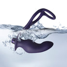 An action photograph of a dark purple silicone prostate massager partially submerged in clear, splashing water against a white background. The wavy water surface splits the device, with the smooth, curved main bulbous arm resting fully underwater amidst small air bubbles. The textured lower section and its open dual-ring loop handle extend out above the waterline, highlighting the toy's waterproof design