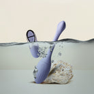 A lavender purple underwater NIYA N1 with a matching remote, partially submerged in water next to a white rock, set against a clean beige background. waterproof design