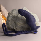 A close-up photograph of a dark blue silicone prostate and perineum massager covered in fine water droplets. The device features a textured, curved bulbous upper head connected to a flat, nubbed base that extends into an open dual-ring loop structure. It rests horizontally on a smooth gray surface in front of a jagged, light gray rock background