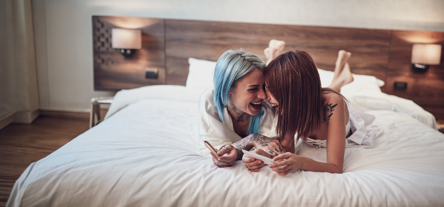 Two women lying on a bed holding a bullet vibrator in a bedroom setting.