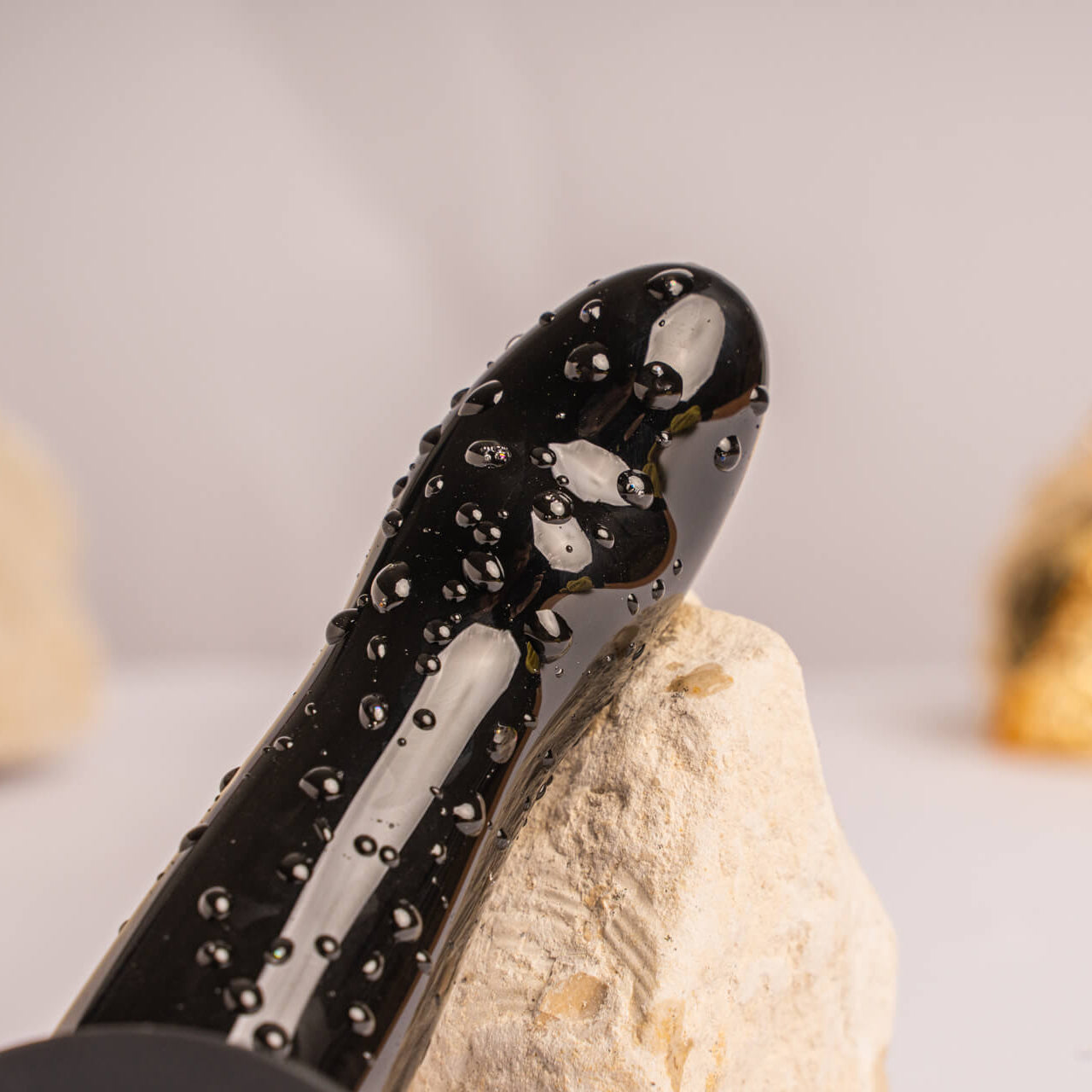 Close up of quest prostate massager, leant on a rock for scale and beaded in drops of water to show it is waterproof